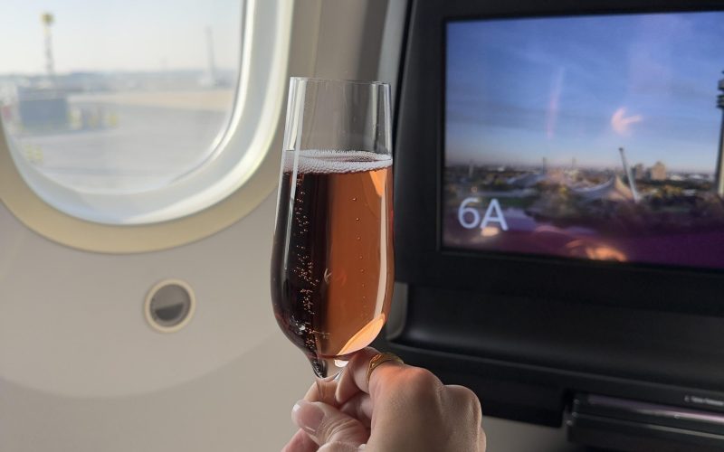 Rosé-Champagner im Glas vor einem Flugzeugfenster bei Sonnenschein – luxuriöser Reisemoment