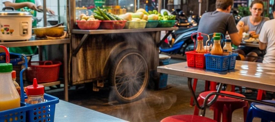 Photorealistic Pad Pak Ruam Mit on a simple table in the foreground of a busy Bangkok night street-food restaurant with neon light, plastic chairs, open wok kitchen, and authentic local atmosphere.